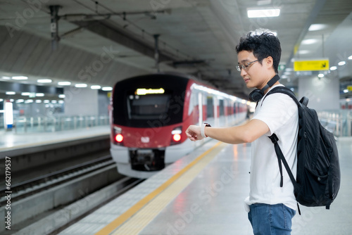Young Asian millenael man standing in train  while looking at smart watch when the train is coming with black backpack and headphone with happiness.