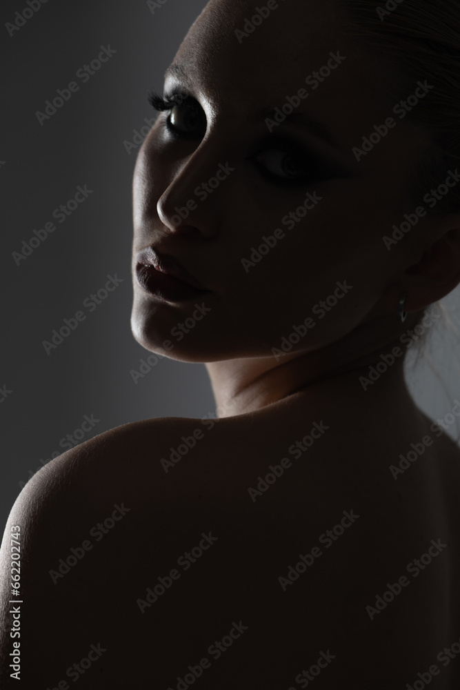 Beauty, fashion, style and make-up concept. Beautiful woman dark silhouette portrait. Model looking with seductive look through her shoulder. Studio shot on gray background