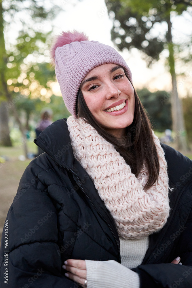 Vertical portrait of a beautiful caucasian young woman looking at camera with a sweet smile wearing warm clothing, standing outdoors in a public park. Vertical portrait of a beautiful caucasian young woman looking at camera with a sweet smile wearing warm clothing, standing outdoors in a public park.