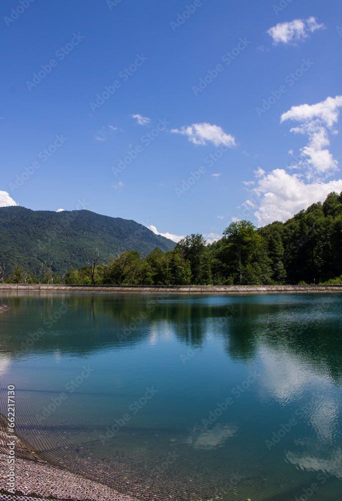 Beautiful mountain landscape - artificial emerald lake with reflection and clear water among hilly shores with green forest on a sunny summer day in Krasnaya Polyana russia
