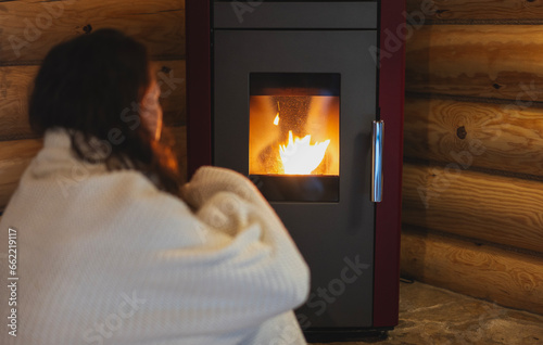 Obraz na płótnie A young woman wraps herself in a warm blanket while sitting by a pellet stove in a wooden log house. Warm home in cold winter concept w salonie