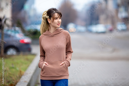 Close portrait of a beautiful slim sexy caucasian brunette girl with dreadlocks in rose pink hoodie dress standing amidst the street and dreamily looking right © Michael Rekochinsky