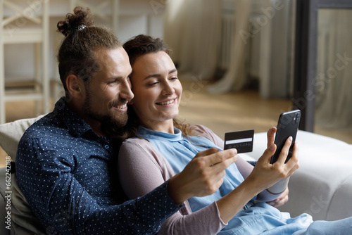 Young married couple of happy customers using smartphone and credit card for online payments from home, shopping on internet with virtual ecommerce app, service, making purchases