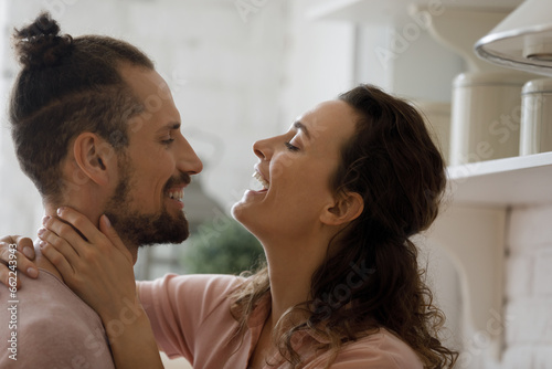 Cheerful excited young dating couple hugging with love, joy, enjoying romantic leisure time together at home. Boyfriend and girlfriend talking, laughing in kitchen domestic interior