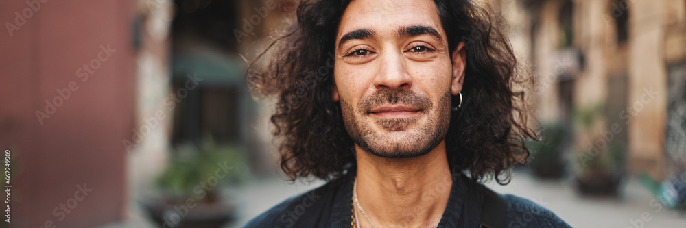 Fototapeta premium Young attractive italian guy with long curly hair and stubble looks into the camera and smiles at old buildings background. Stylish man with an earring in his ear and lot of chains