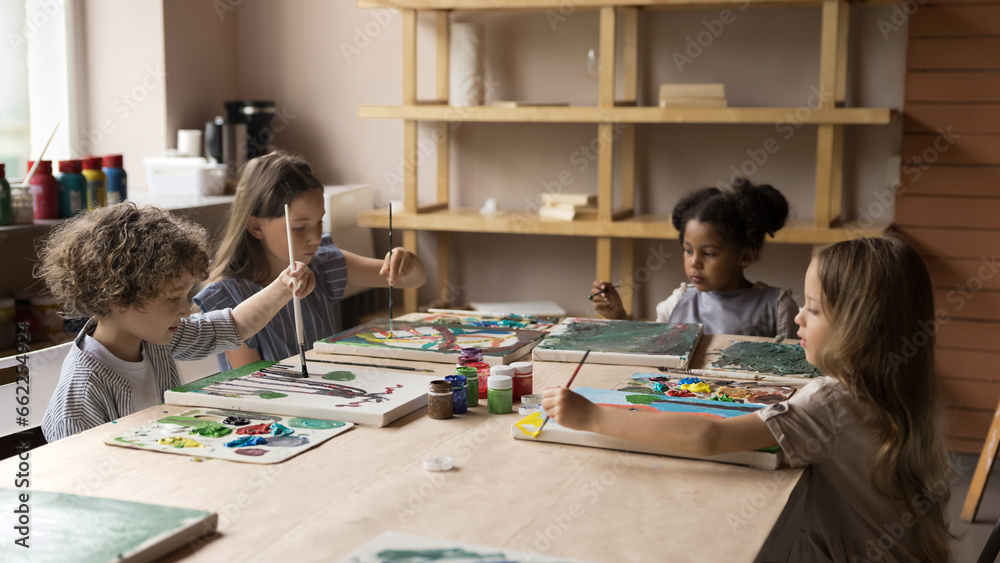 Fototapeta premium Group of cute little serious boy and multiethnic girls sit at table in art-studio engaged in artistic class, painting pictures with paints on canvas. Children development, creative hobby, day-care