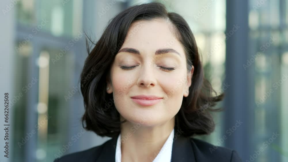 Close up. Happy young beautiful businesswoman relaxing with closed eyes standing on the street near the office building. A smiling joyful brunette woman in a formal suit breathes deeply and enjoys Close up. Happy young beautiful businesswoman relaxing with closed eyes standing on the street near the office building. A smiling joyful brunette woman in a formal suit breathes deeply and enjoys