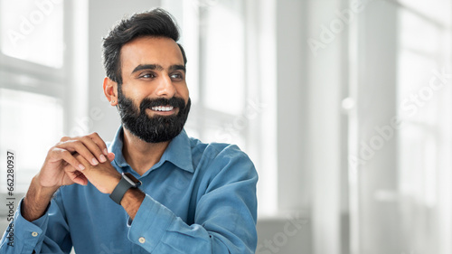 Confident smiling bearded indian man posing at office, looking aside at copy space, sitting at workplace, panorama