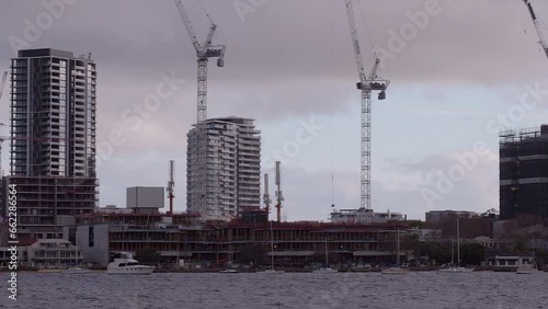 Grey cloudy sky frames riverside building constructions Cranes overhead. Pan across skyscrapers in background with houses and boats in foreground