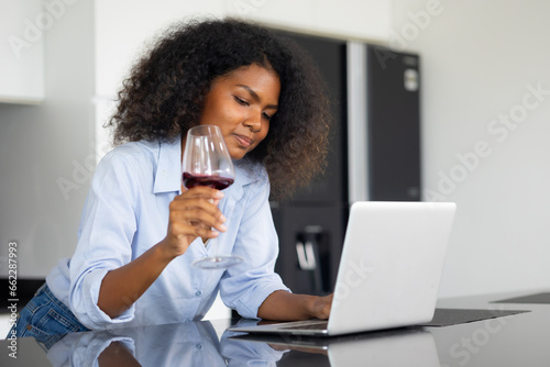 Enjoy drinking wine. African-American female working at kitchen table in front of generic laptop computer, online banking app for paying bills