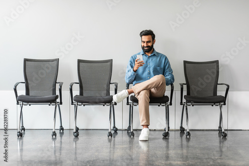 Businessman sitting in recruitment waiting room for interview with HR, using cellphone, full length