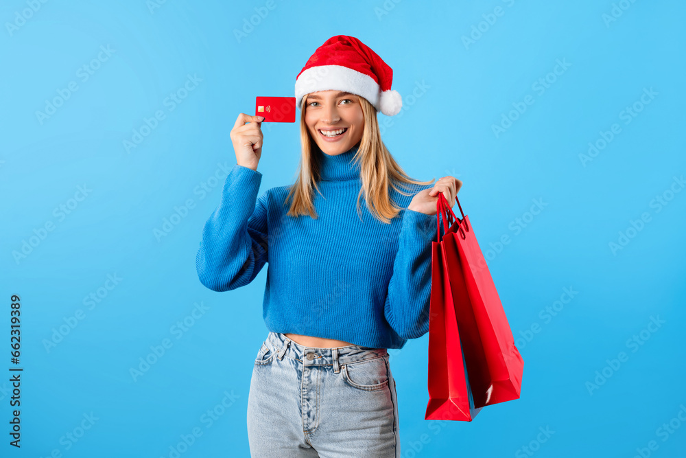 Online Christmas sale. Happy lady in Santa hat holding shopping bags and credit card, posing on blue studio background