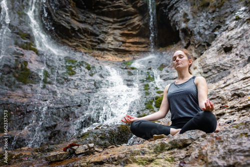 Young woman practicing yoga  sitting in om pose on the rock near the beautiful waterfall. Relax  nature  and wellbeing concepts.