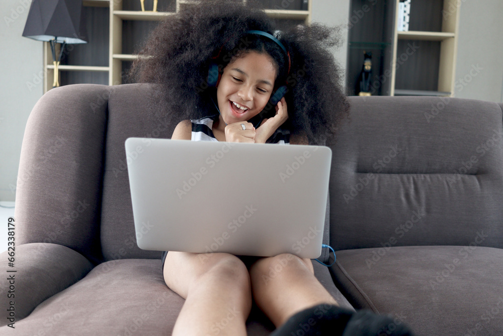 Naklejka premium Happy smiling beautiful teenage girl with curly hair wearing headphones, using laptop computer for practicing to sing song while sitting on sofa in home living room.