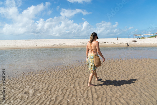 A beautiful woman in a bikini walking on the wavy sand of a beach. Blue sky and clouds in the background. Taquari Beach in Valenca, Brazil.