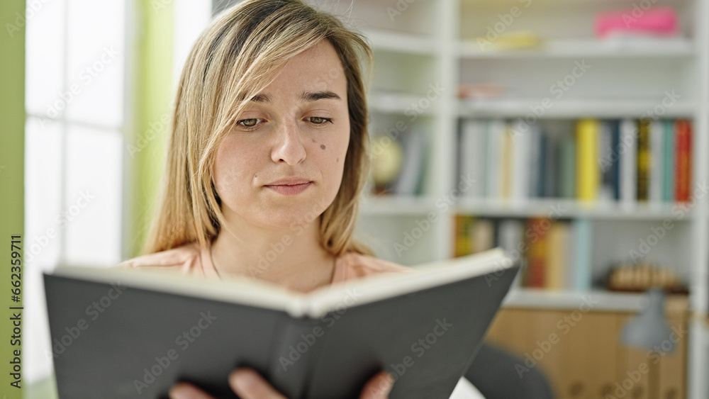 Young blonde woman student reading book sitting on table at library university Young blonde woman student reading book sitting on table at library university