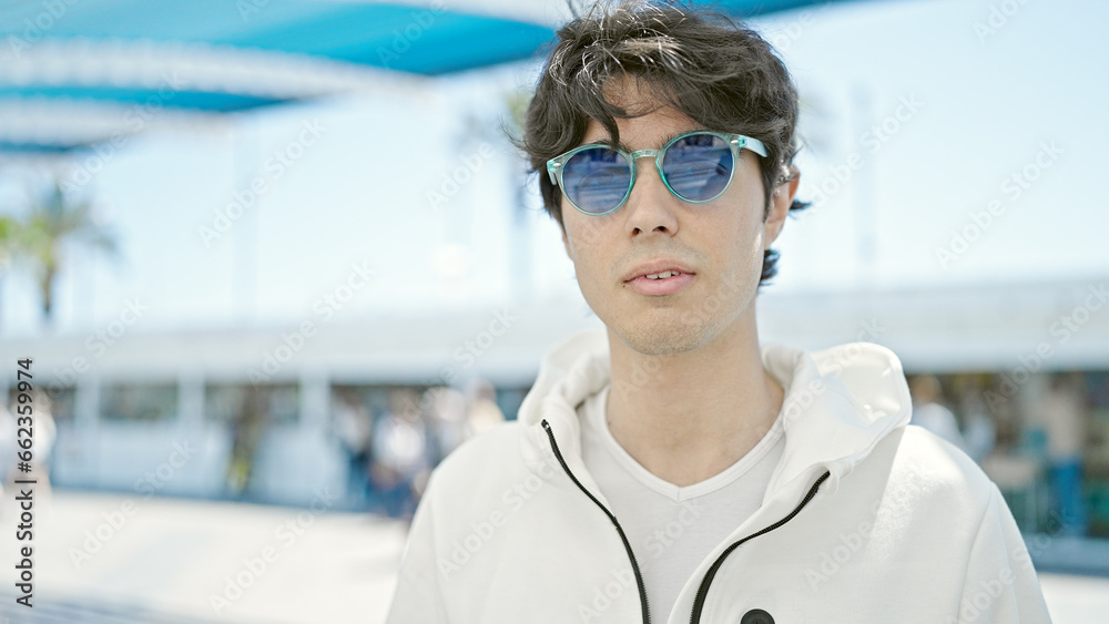 Young hispanic man standing with serious expression wearing sunglasses at park