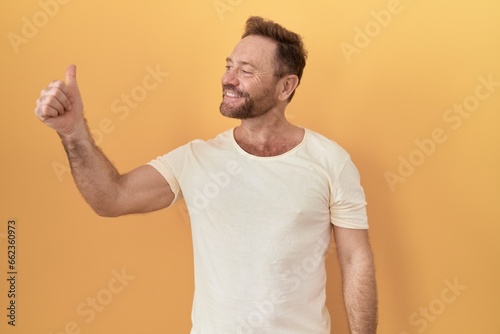 Middle age man with beard standing over yellow background looking proud, smiling doing thumbs up gesture to the side