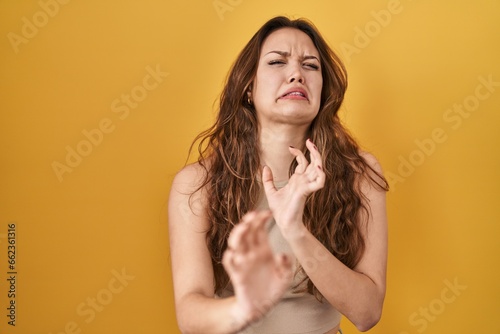 Young hispanic woman standing over yellow background disgusted expression, displeased and fearful doing disgust face because aversion reaction. with hands raised photo