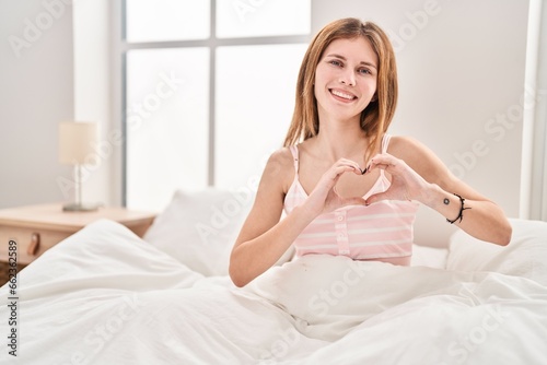 Young blonde woman doing heart gesture sitting on bed at bedroom