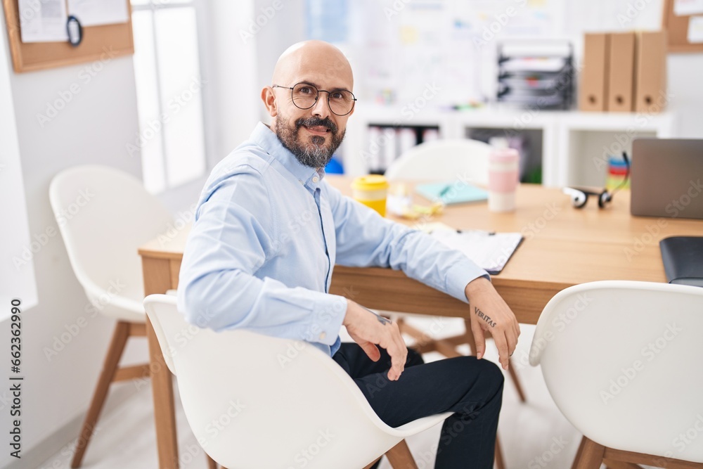 Young bald man business worker smiling confident sitting on table at office