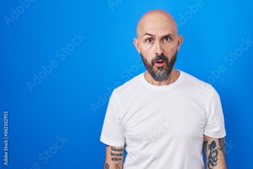 Hispanic man with tattoos standing over blue background in shock face, looking skeptical and sarcastic, surprised with open mouth