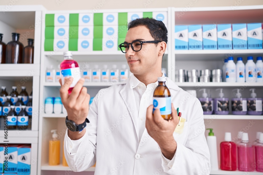 Young hispanic man pharmacist smiling confident holding medication bottles at pharmacy