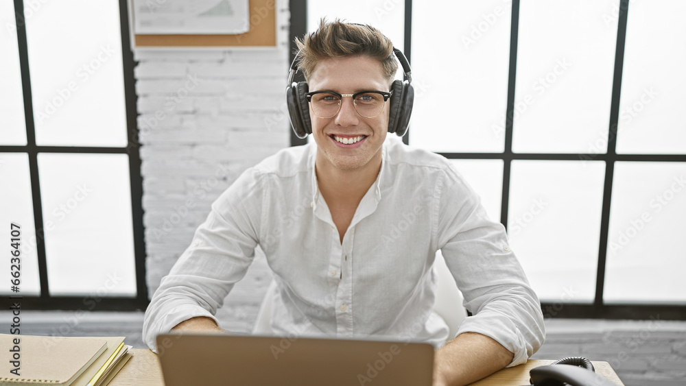 Smiling young caucasian business worker enjoying his job, confidently managing work online on laptop, with headphones on his desk at the office.