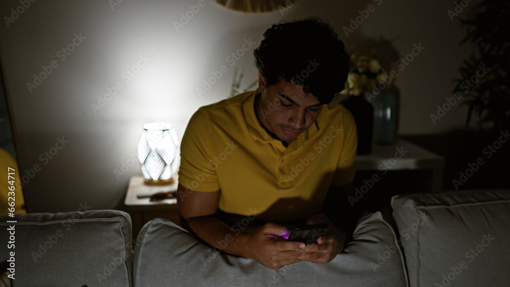 Young latin man using smartphone leaning on sofa at home