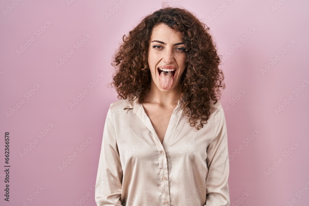Hispanic woman with curly hair standing over pink background sticking tongue out happy with funny expression. emotion concept. Hispanic woman with curly hair standing over pink background sticking tongue out happy with funny expression. emotion concept.