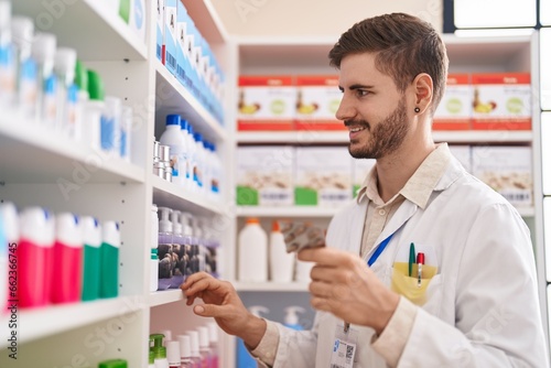 Young caucasian man pharmacist holding pills of shelving at pharmacy