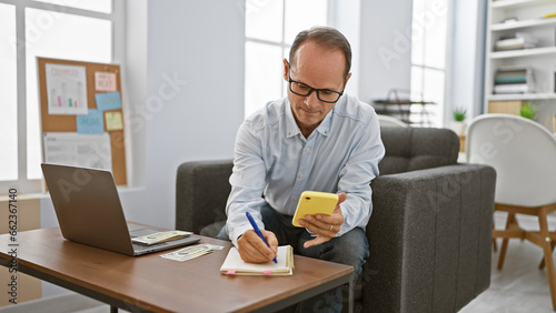 Handsome middle age business man, a diligent worker at his office, engrossed in taking meticulous notes on his smartphone amid an elegant room setting, portraying professional success.