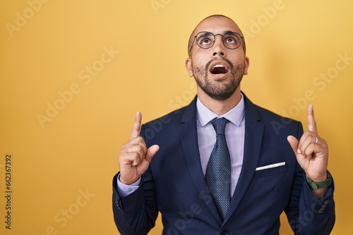 Hispanic man with beard wearing suit and tie amazed and surprised looking up and pointing with fingers and raised arms.