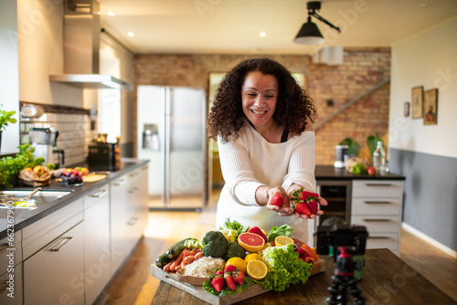 Middle aged female mixed ethnicity food nutritionist and blogger recording a video about a healthy recipe with organic fruits and vegetables in her kitchen at home photo