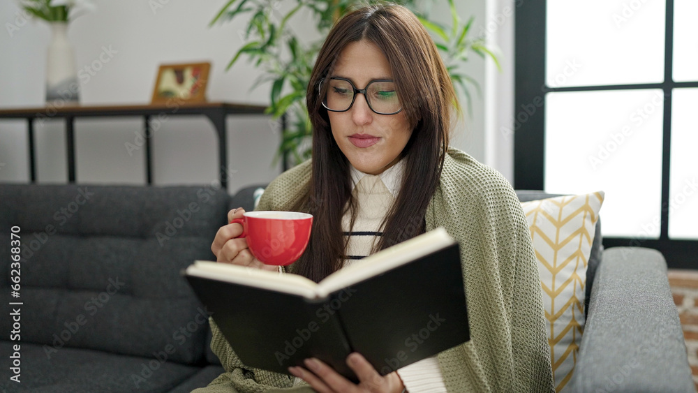 Young beautiful hispanic woman reading book and drinking coffee sitting on sofa at home Young beautiful hispanic woman reading book and drinking coffee sitting on sofa at home