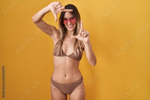 Young hispanic woman wearing bikini over yellow background smiling making frame with hands and fingers with happy face. creativity and photography concept.