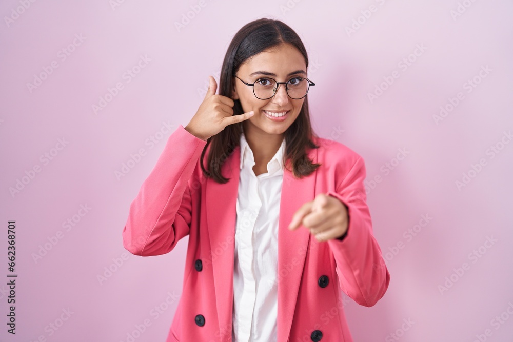 Young hispanic woman wearing business clothes and glasses smiling doing talking on the telephone gesture and pointing to you. call me.