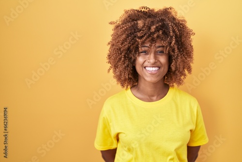 Young hispanic woman with curly hair standing over yellow background with a happy and cool smile on face. lucky person.