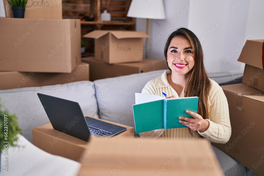 Young beautiful hispanic woman using laptop writing on notebook at new home Young beautiful hispanic woman using laptop writing on notebook at new home