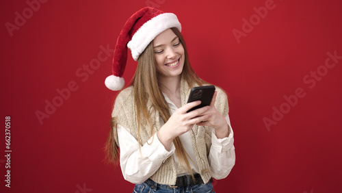 Young blonde woman wearing christmas hat using smartphone over isolated red background