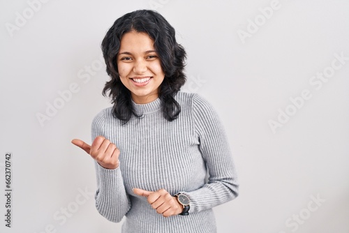 Hispanic woman with dark hair standing over isolated background pointing to the back behind with hand and thumbs up, smiling confident