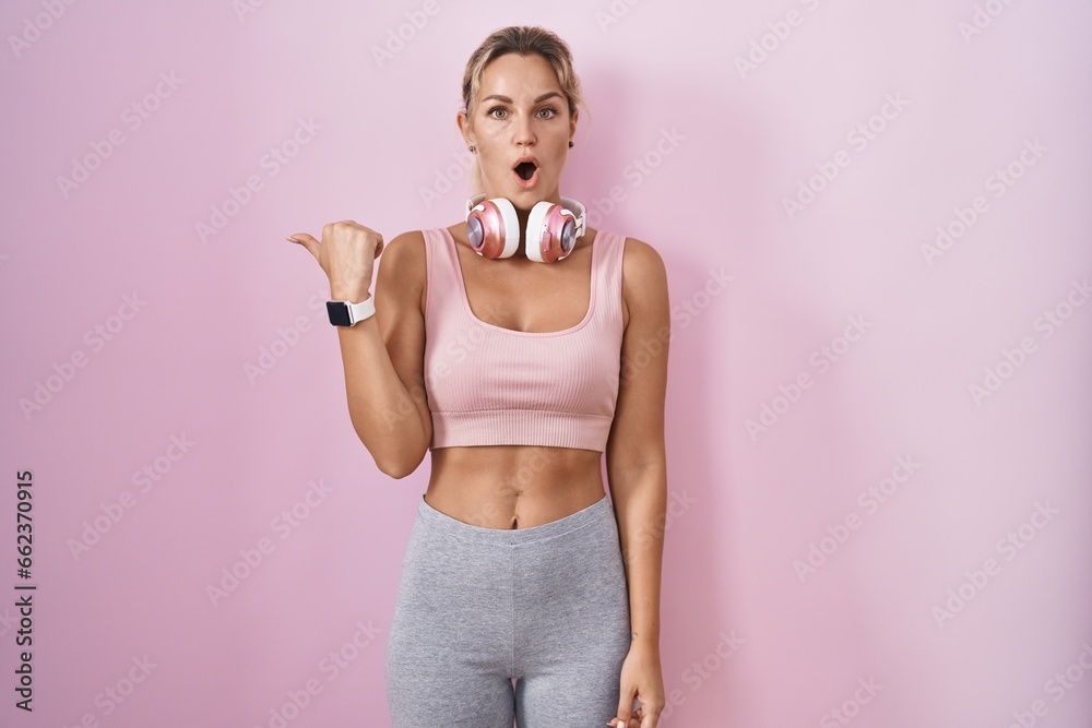 Young blonde woman wearing sportswear and headphones surprised pointing with hand finger to the side, open mouth amazed expression. Young blonde woman wearing sportswear and headphones surprised pointing with hand finger to the side, open mouth amazed expression.