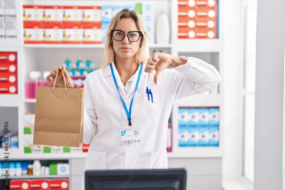 Young blonde woman working at pharmacy drugstore holding paper bag with angry face, negative sign showing dislike with thumbs down, rejection concept Young blonde woman working at pharmacy drugstore holding paper bag with angry face, negative sign showing dislike with thumbs down, rejection concept