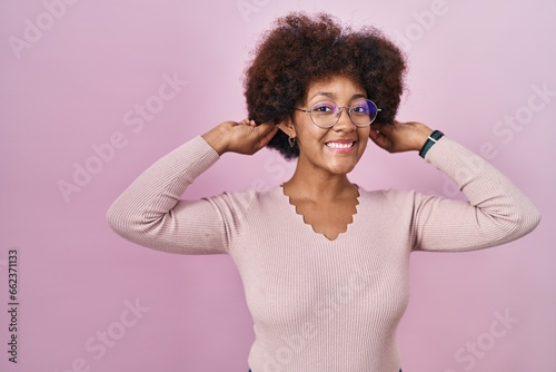 Young african american woman standing over pink background smiling pulling ears with fingers  funny gesture. audition problem