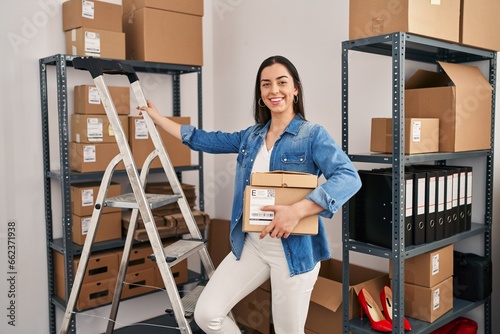 Young beautiful hispanic woman ecommerce business worker standng on ladder holding packages at office photo