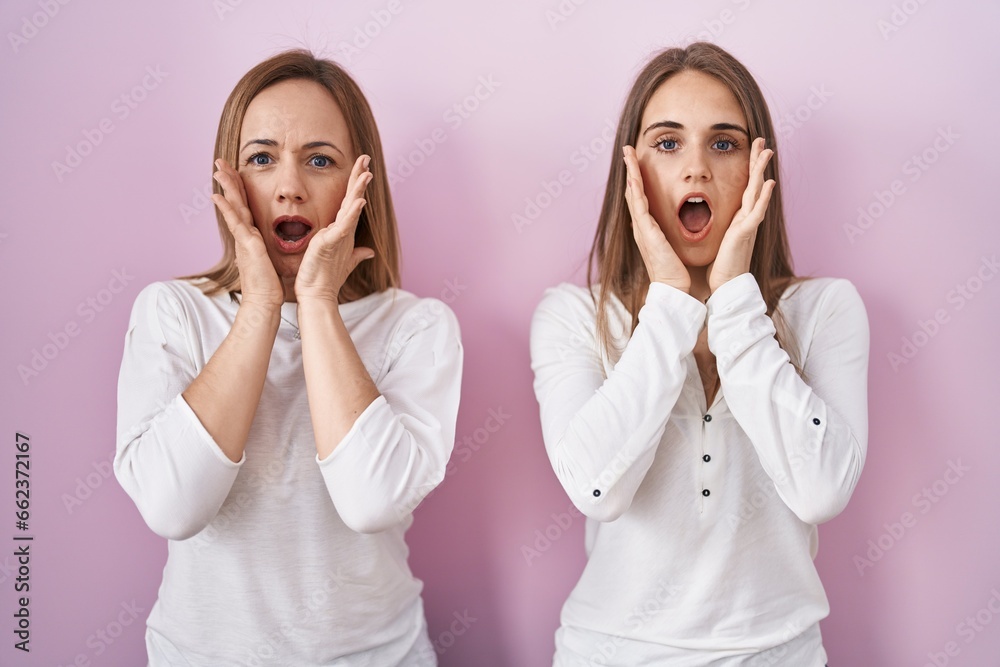 Middle age mother and young daughter standing over pink background afraid and shocked, surprise and amazed expression with hands on face
