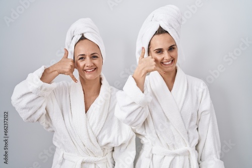 Middle age woman and daughter wearing white bathrobe and towel smiling doing phone gesture with hand and fingers like talking on the telephone. communicating concepts.