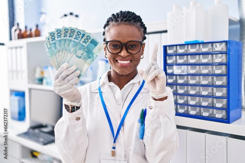 African woman with braids working at scientist laboratory holding money smiling with an idea or question pointing finger with happy face  number one