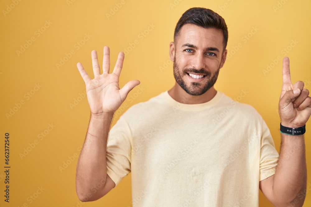 Handsome hispanic man standing over yellow background showing and pointing up with fingers number six while smiling confident and happy. Handsome hispanic man standing over yellow background showing and pointing up with fingers number six while smiling confident and happy.