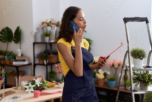 Young african american woman florist talking on smartphone reading document at flower shop